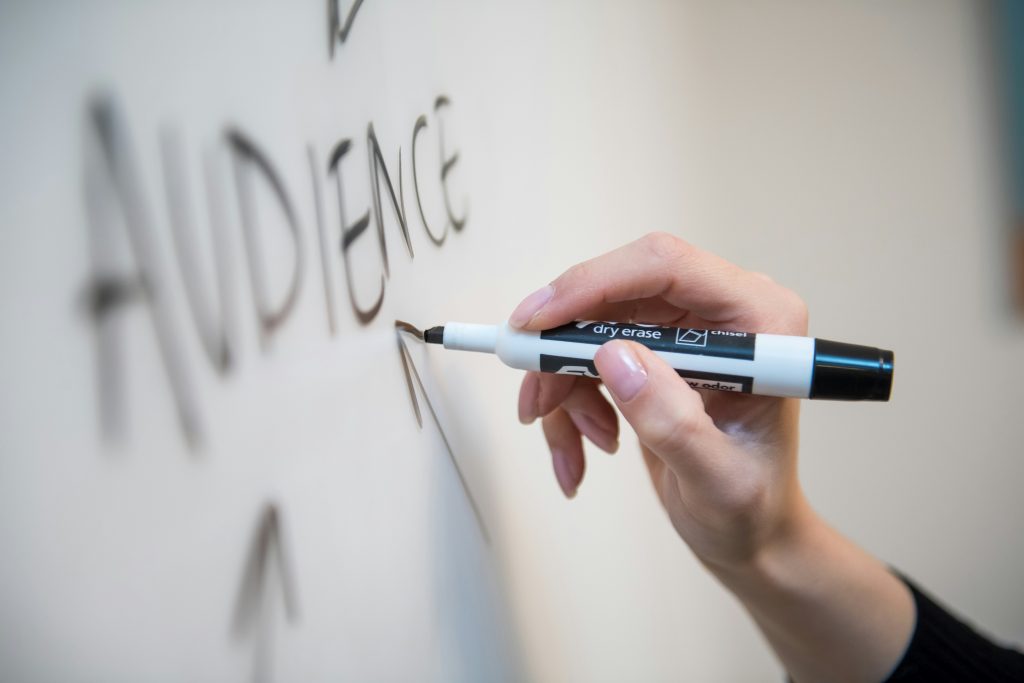 Close-up picture a female hand holding a pen, drawing an arrow over a whiteboard pointing the the word "audience"